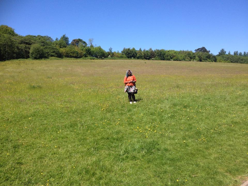 picture of a tiny me wearing an orange top, in the middle of a green grassy field with little yellow and purple wildflowers. there are trees in the background and a blue sky